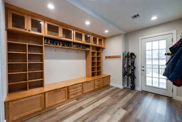 Mudroom with extensive wooden storage: upper cabinets, open shelves, shoe cubbies, drawers, and hooks; skis lean by the door nearby.